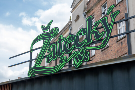 Žateck&yacute; beer brand sign, black cursive lettering with green neon outline on metal frame above corrugated facade, historic brick and plaster buildings, blue sky, editorial