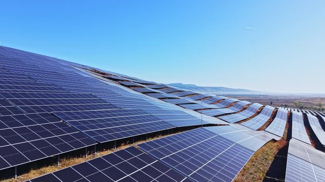 Rows of solar panels cover a hillside, capturing sunlight for energy. The clear sky shows no clouds, indicating a sunny day. The panels are arranged in waves along the slope.