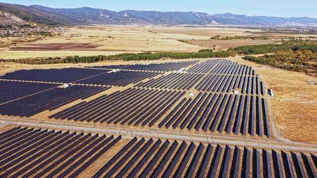 Solar panels cover a large area in a flat landscape. The scene shows rows of solar panels aligned in the field with distant mountains and trees.