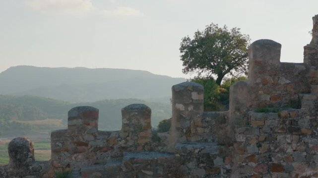 Sunlit ancient stone battlement with solitary oak tree and layered mountain ridges in soft afternoon haze, warm textured masonry overlooking peaceful valley vista