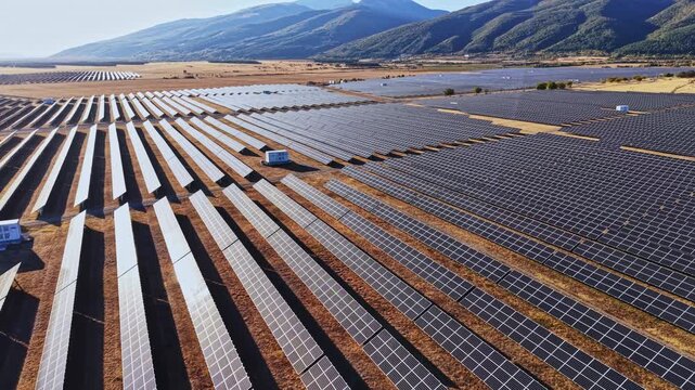 Wide view of a solar panel field located in a rural area surrounded by mountains. The panels are aligned in rows under clear skies during the day.