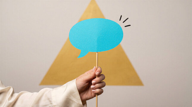Hand holding a blue speech bubble cutout on a stick in front of a yellow triangle on a wall