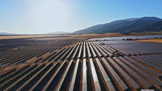 Rows of solar panels are aligned neatly in a renewable energy farm located in a mountainous area. The sun is shining brightly and casts clear shadows.