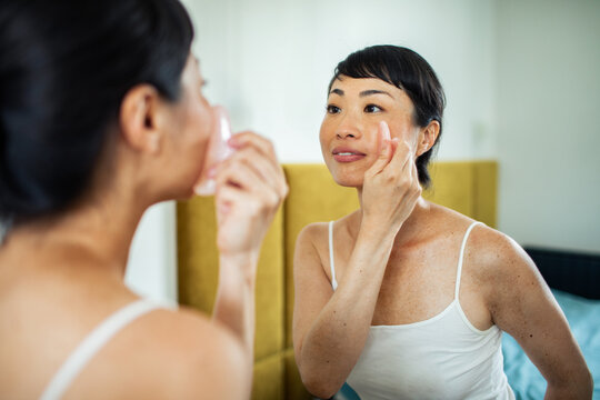 Woman using gua sha and applying eye cream at mirror