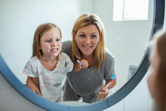 Mother helping daughter brush teeth at bathroom mirror