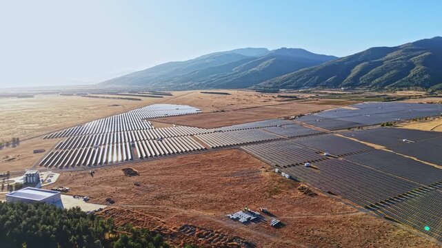 Sunlight shines on a vast solar panel field in a valley with mountains in the background. The panels are arranged in neat rows across the landscape.