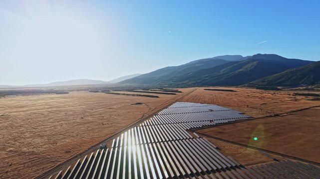 Rows of solar panels are set up in a wide desert area with mountains in the background. The sun shines brightly, creating reflections on the panels.