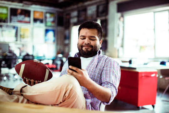 Man checking smartphone while holding football in office