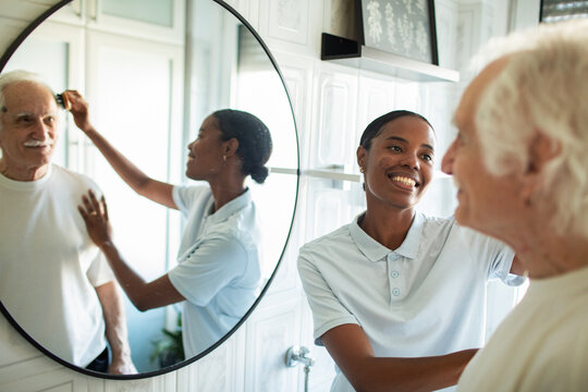 Caregiver brushing elderly man's hair in bathroom mirror
