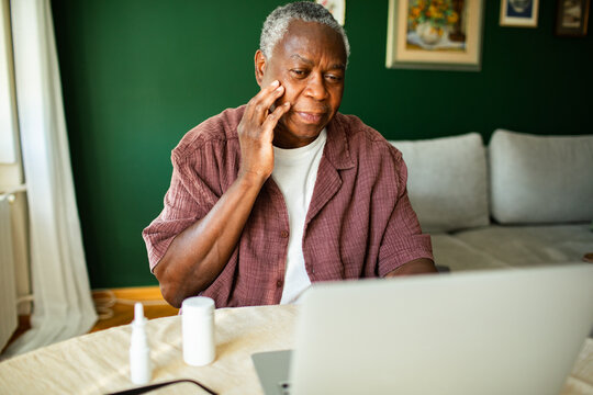 Senior man touching cheek while using laptop at home