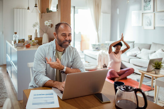 Man on video call at home while partner does yoga