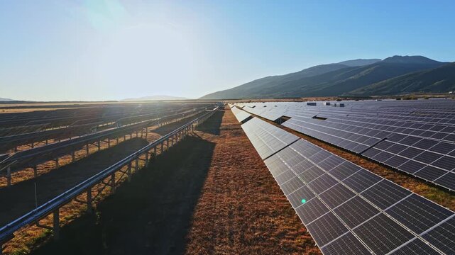 Rows of solar panels stretch across a valley under a bright sun. Mountains are visible in the distance, and the scene takes place in the afternoon.