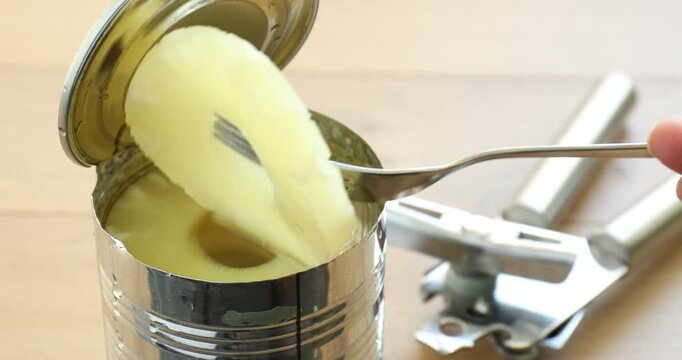 Woman taking pickled pineapple ring from tin and can opener at wooden table, closeup