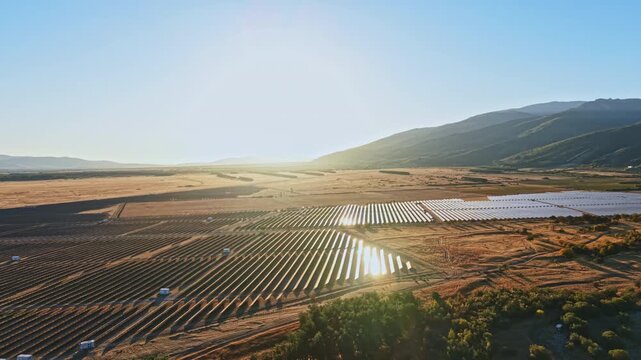 Solar panels are arranged in rows across a field in a rural area. Mountains rise in the background under a clear blue sky. Sunlight reflects off the panels.