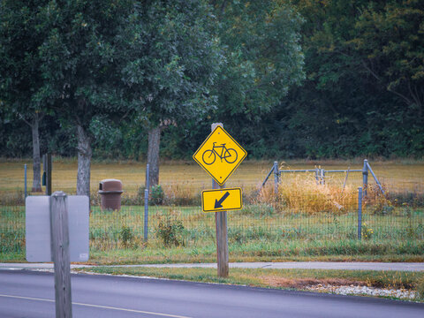 Yellow bike crossing warning sign with directional arrow posted along a paved park road and bicycle trail