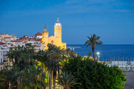 The illuminated Church of Sant Bartomeu and Santa Tecla rises above the old town and Mediterranean Sea in the coastal town of Sitges, Spain, Catalonia region.