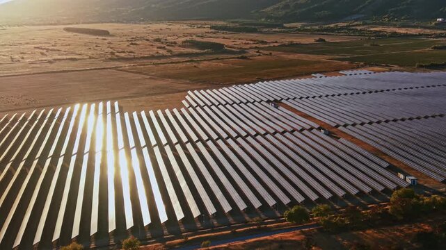 Rows of solar panels spread out across a countryside area. The sun sets in the background, creating reflections on the panels.