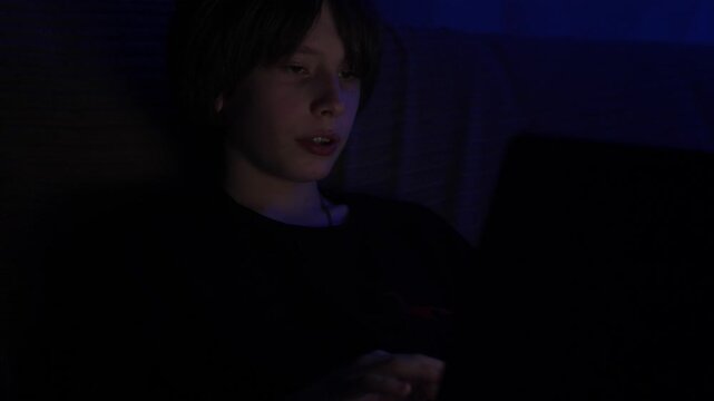 A young teenage boy sits in a dark room at night, deeply focused while working on his laptop. The cool blue light of the screen brightly illuminates his face as he types