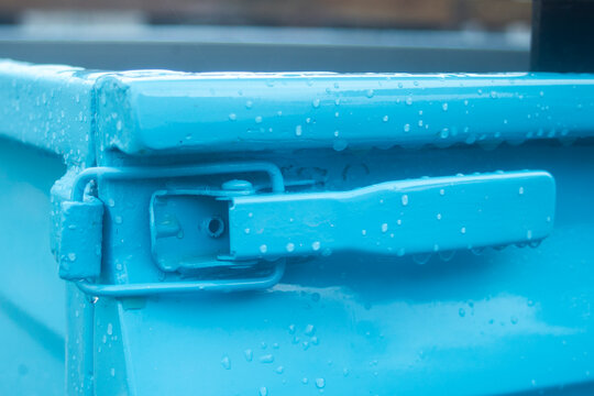 Close up of a blue metal truck bed tailgate latch covered in raindrops.
A high-quality close-up photograph focusing on a heavy-duty toggle latch mechanism on the tailgate corner of a vintage pickup 