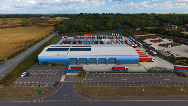 Aerial view of an industrial logistics warehouse facility with rows of parked HGVs and trailers in a paved yard under a blue sky in Bury St Edmunds, England, United Kingdom.