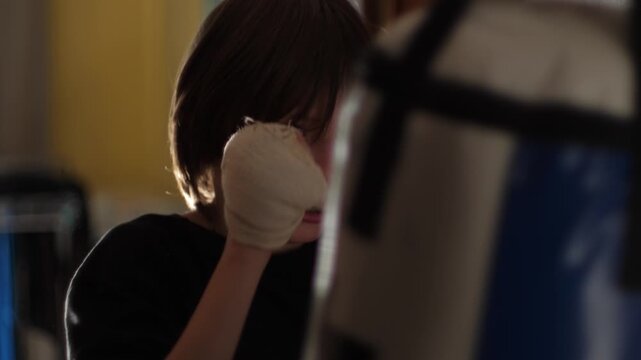 Young boy in a black t-shirt and hand wraps actively training at home, practicing punches on a punching bag. Child athlete working out diligently in a room, developing strength and endurance.