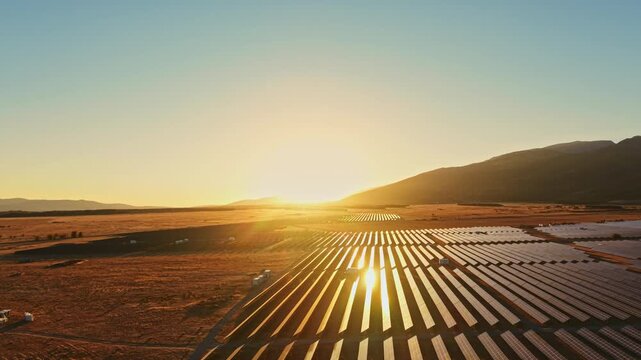 Sunlight shines on rows of solar panels in a valley during sunset. The landscape shows mountains in the distance. This scene represents renewable energy and technology.