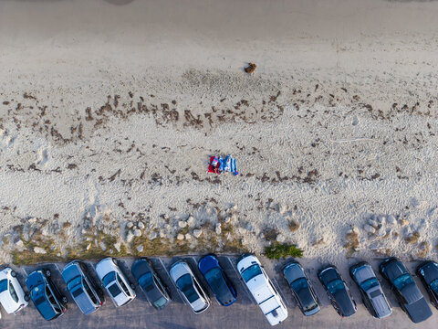 Aerial view of the waterfront showing a row of parked cars along the sandy beach with scattered seaweed and people relaxing on a blanket in Santa Barbara, California, United States.
