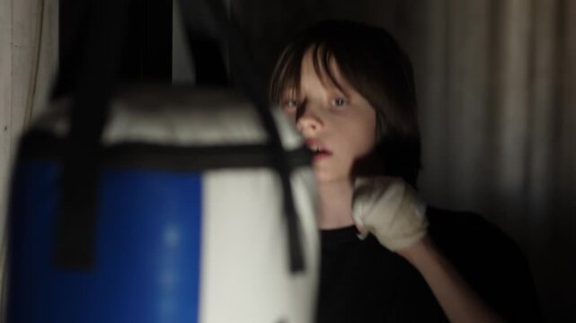 A young boy intensely trains by punching a heavy bag in a dim room. He wears hand wraps, showing fierce determination, focus, and a strong passion for martial arts and fitness.