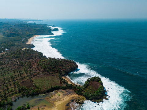 Aerial view of Pantai Wonogoro featuring rugged cliffs, lush green vegetation, and white waves crashing against the sandy shoreline in Malang, East Java, Indonesia.