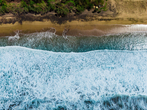 Aerial view of Pantai Wonogoro with turquoise waves crashing onto the sandy shore and lush green trees along the coastline in Malang, East Java, Indonesia.