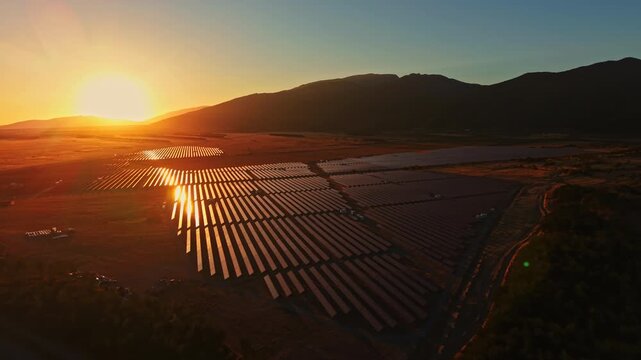 The sun sets over a solar farm, casting long shadows across rows of solar panels in a field. Mountains rise in the background as light reflects off the panels.