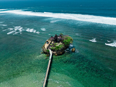 Aerial view of Balekambang Beach temple on a small rocky island connected by a long bridge to the shore, surrounded by clear turquoise water and waves in Malang, East Java, Indonesia.