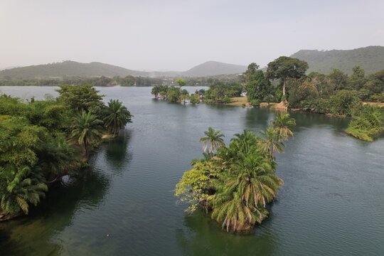 Aerial view of the Volta River featuring lush green islands with palm trees, calm dark water, and rolling hills in the background in Akosombo, Eastern Region, Ghana.