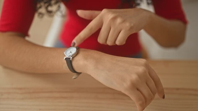 Young woman points finger to watch on wrist while sitting at wooden dining table in building kitchen; impatience hurry.