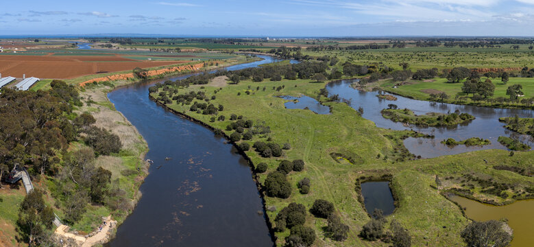 Aerial drone view following the Werribee River in Werribee South, Melbourne, Victoria, Australia, with flooded river water winding between eroded banks, eucalyptus bushland, and ploughed farmland.