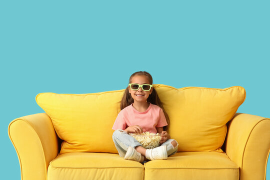Entertainment and leisure, happy little girl wearing 3d glasses eating popcorn while sitting on a yellow sofa against a blue background