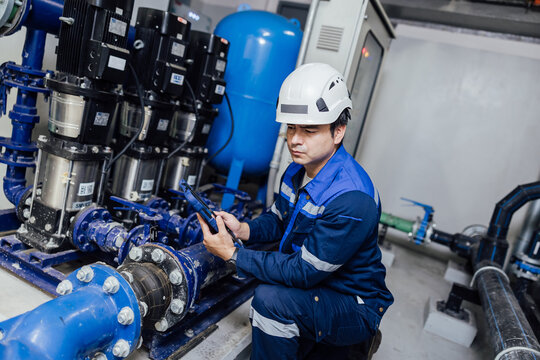 Industrial technician in safety gear servicing a blue pump system inside a mechanical room, inspecting valves and pipes with a wrench during maintenance at a utility facility.
