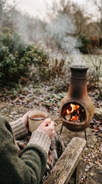 Person sitting in a wooden chair outdoors wrapped in a blanket holding a warm mug near a burning clay chimenea in a garden