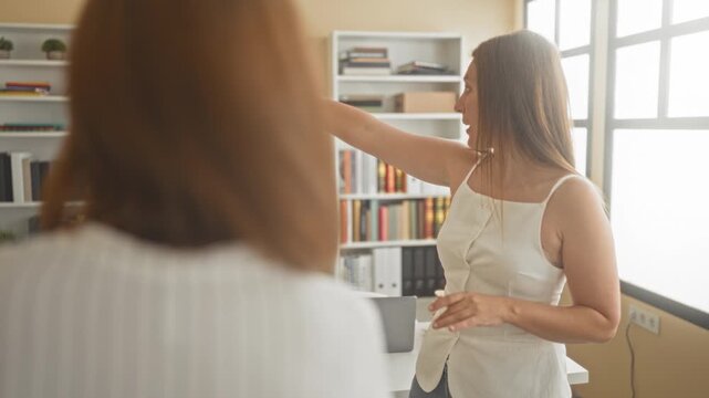 Woman pointing arm toward a bright window while speaking to a colleague in an office building; concern discussion.