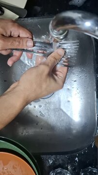 Close-up of hands rinsing metal forks under running water in a stainless steel kitchen sink, documenting an everyday dishwashing routine in a home kitchen