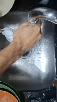 Close-up of a person rinsing metal spoons under running water in a stainless steel kitchen sink, illustrating a simple household cleaning routine after a meal