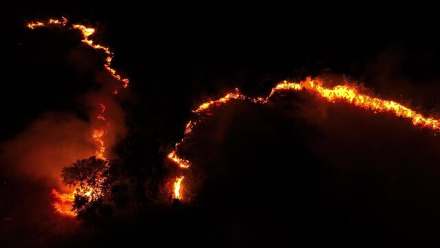 Drone view at night capturing a destructive wildfire moving across hills, with intense flames and heavy smoke illustrating the growing impact of climate change and worsening air quality worldwide. 4k.