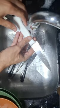 Person carefully rinses kitchen knives under running water in a stainless steel sink, showing a close-up of a common kitchen cleaning task and careful utensil handling