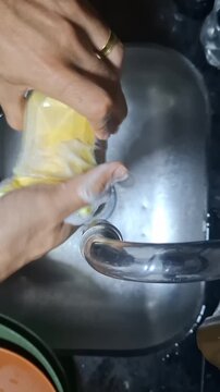 Timelapse video of a person washing a drinking glass and cutlery with a yellow sponge and dish soap in a stainless steel kitchen sink, showing an everyday household cleaning routine