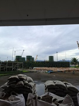 Bulk construction materials stored at an outdoor worksite, with stacked filled sacks in the foreground and industrial tanks and cranes in the background under an overcast sky