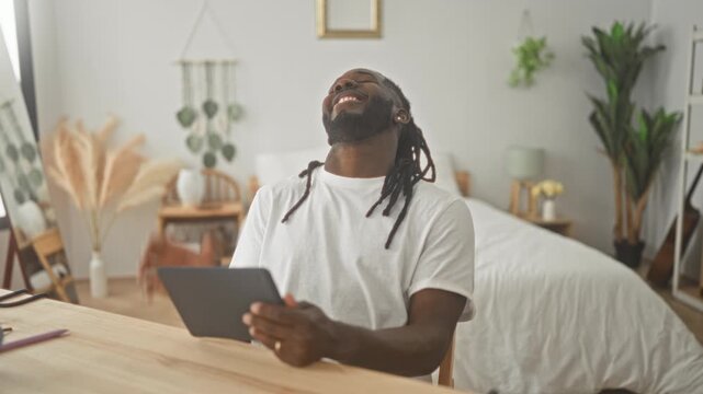Man holding tablet and tapping screen, laughing with hand to face and smiling at device in tidy bedroom setting; relaxation joy.
