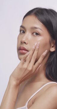 Asian woman using serum dropper on cheek and smiling at camera. She gently taps her skin in studio.