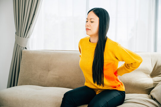 Asian woman holds her lower back in unbearable pain on a sofa