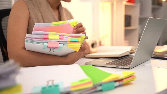 Woman Carrying Stack of Papers: A woman sits at her desk, holding a large stack of documents and folders, with a laptop and notebook in front of her.