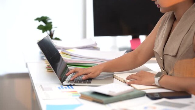 Focused Professional at Work: A woman meticulously works on her laptop at a desk piled high with documents and charts, surrounded by the organized chaos of a busy office environment.